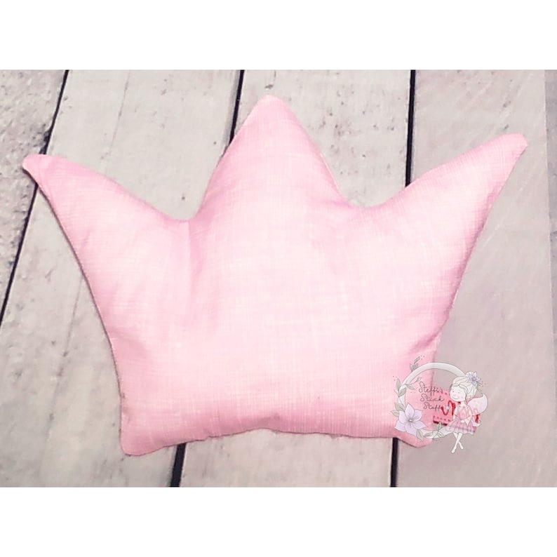May include: A pink crown-shaped pillow made of soft fabric. The pillow is on a light wood surface.