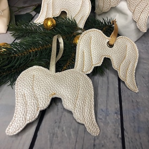 May include: White leather angel wings with gold stitching, hanging from a brown ribbon. The wings are a Christmas ornament.