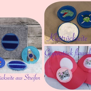 Pode incluir: Um conjunto de patches de feltro com v&aacute;rios designs, incluindo um foguete, um planeta, um dinossauro, um ninja e gatos. Os patches t&ecirc;m um suporte de velcro para fixar em tecido. O texto "Klett R&uuml;ckseite" e "Gegenst&uuml;ck flauschig" &eacute; vis&iacute;vel na imagem.