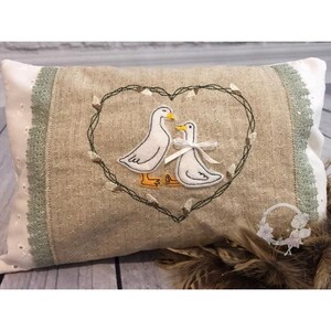 Op de afbeelding: Een beige kussen met een groen geborduurd hartontwerp met twee witte eenden. Het kussen heeft een witte kantversiering met groene accenten.