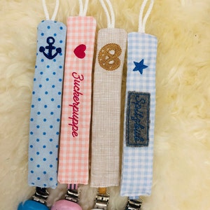 May include: Four colorful fabric baby teethers with different embroidered designs and wooden clips. The designs include an anchor, a heart, a pretzel, and a star. The teethers are attached to wooden clips with different colored beads.