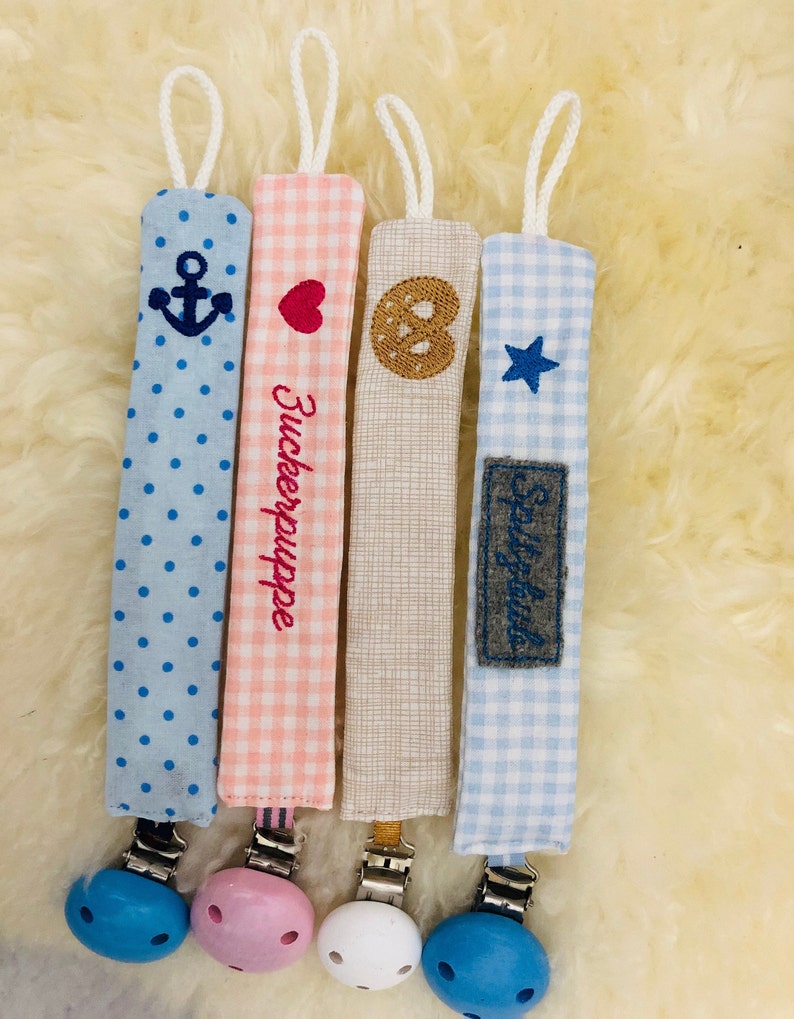 May include: Four colorful fabric baby teethers with different embroidered designs and wooden clips. The designs include an anchor, a heart, a pretzel, and a star. The teethers are attached to wooden clips with different colored beads.