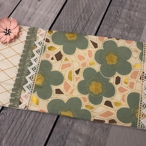 Può includere: Una piccola pochette rettangolare con un motivo floreale in tonalità di verde, rosa e oro. La pochette è rifinita con un bordo in pizzo bianco e un fiore di stoffa rosa.
