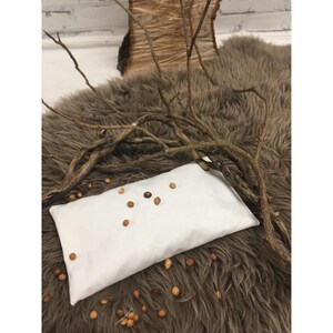 May include: White fabric eye pillow filled with small brown seeds, resting on a brown faux fur surface with a branch of twigs on top.