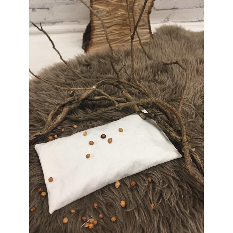 May include: White fabric eye pillow filled with small brown seeds, resting on a brown faux fur surface with a branch of twigs on top.
