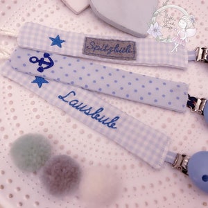 May include: Three blue and white fabric baby teethers with a star, anchor, and the word "Lausbub" embroidered on them. Each teether has a blue wooden clip.