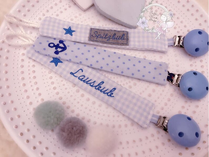 May include: Three blue and white fabric baby teethers with a star, anchor, and the word "Lausbub" embroidered on them. Each teether has a blue wooden clip.