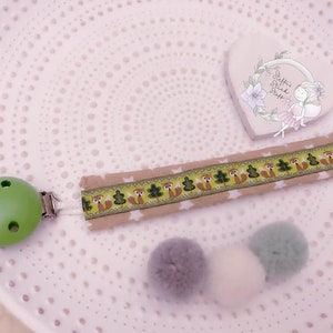 May include: A green wooden baby teether with a clip attached to a beige fabric ribbon with a repeating pattern of foxes, trees, and stars. The ribbon is attached to three pom-poms in shades of gray and white. The text "Steffis Gl&uuml;ck St&uuml;ck" is visible on a heart-shaped decoration in the background.