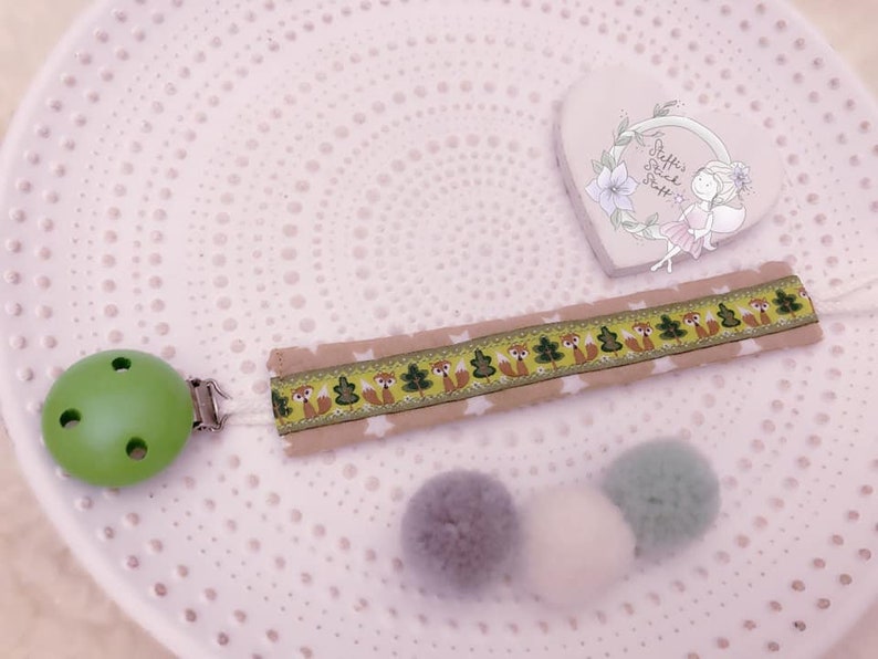 May include: A green wooden baby teether with a clip attached to a beige fabric ribbon with a repeating pattern of foxes, trees, and stars. The ribbon is attached to three pom-poms in shades of gray and white. The text "Steffis Gl&uuml;ck St&uuml;ck" is visible on a heart-shaped decoration in the background.