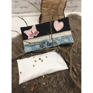 May include: A white fabric eye pillow filled with brown seeds. The pillow is lying on a brown fluffy surface. A black and blue fabric pillow with a pink butterfly and heart applique is in the background. The pillow has white lace trim and the name "Flora" embroidered on it.