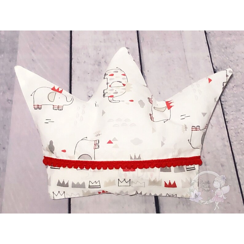 May include: A white crown-shaped pillow with a red trim and a pattern of gray elephants and crowns. The pillow is made of fabric and has a soft, plush texture.