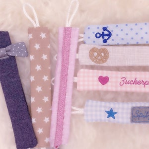 May include: Set of 7 colorful fabric baby teethers with different embroidered designs, including a bow tie, stars, stripes, a pretzel, an anchor, a heart, and a star. The teethers are made of soft fabric and are perfect for babies to chew on.