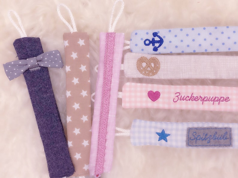 May include: Set of 7 colorful fabric baby teethers with different embroidered designs, including a bow tie, stars, stripes, a pretzel, an anchor, a heart, and a star. The teethers are made of soft fabric and are perfect for babies to chew on.