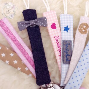 May include: A set of seven colorful fabric pacifier clips with different patterns and embroidered designs. The designs include a bow tie, a heart, a star, a pretzel, an anchor, and two different striped patterns. The clips are attached to a metal clip.