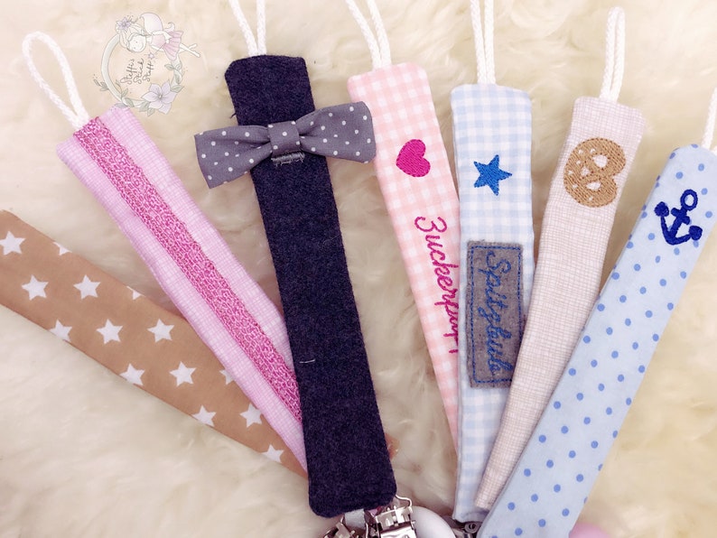 May include: A set of seven colorful fabric pacifier clips with different patterns and embroidered designs. The designs include a bow tie, a heart, a star, a pretzel, an anchor, and two different striped patterns. The clips are attached to a metal clip.