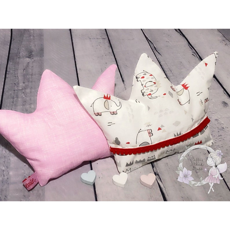 May include: Two decorative pillows in the shape of a crown. The crown pillow is white with a red trim and a pattern of gray elephants. The other pillow is pink and has a star shape. Both pillows are on a wooden surface.