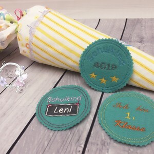 Pode incluir: Tr&ecirc;s c&iacute;rculos de feltro turquesa com costuras douradas e texto bordado. O primeiro c&iacute;rculo diz "Schulkid 2019" com tr&ecirc;s estrelas douradas. O segundo c&iacute;rculo diz "Schulkid Leni" com um quadro negro. O terceiro c&iacute;rculo diz "Ich bin 1. Klasse".