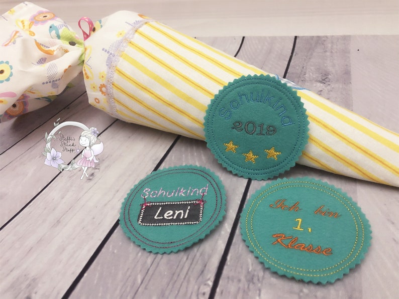 Pode incluir: Tr&ecirc;s c&iacute;rculos de feltro turquesa com costuras douradas e texto bordado. O primeiro c&iacute;rculo diz "Schulkid 2019" com tr&ecirc;s estrelas douradas. O segundo c&iacute;rculo diz "Schulkid Leni" com um quadro negro. O terceiro c&iacute;rculo diz "Ich bin 1. Klasse".