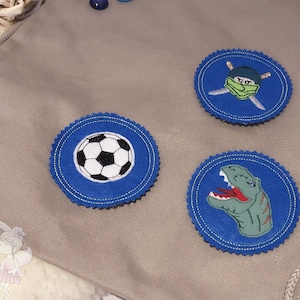 Könnte beinhalten: Drei blaue und weiße Filzpatches mit gestickten Designs. Die Patches zeigen einen Fußball, einen Dinosaurierkopf und einen Ninja mit Schwertern.