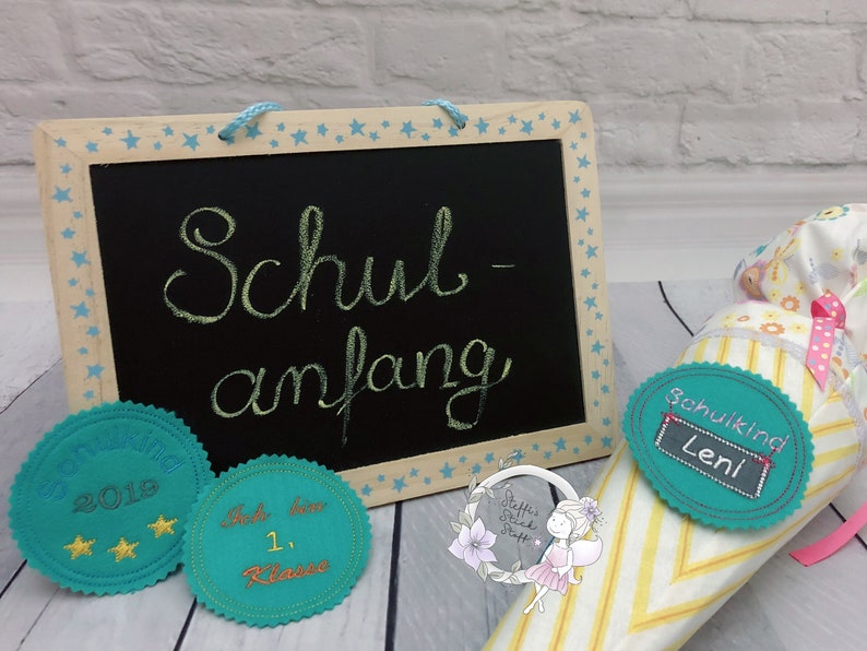 Pode incluir: Um cone escolar de feltro azul turquesa com um padr&atilde;o listrado amarelo e branco. O cone tem uma etiqueta de feltro azul turquesa com o texto "Schulkind Leni". Um pequeno quadro-negro com o texto "Schul-anfang" est&aacute; no fundo.