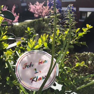 Pode incluir: Um c&iacute;rculo branco com bordado colorido que diz "Ich bin 1. Klasse" em alem&atilde;o. O c&iacute;rculo est&aacute; preso a uma planta verde com flores roxas.