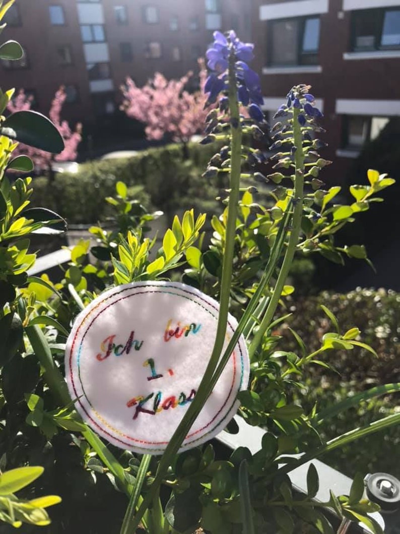 Pode incluir: Um c&iacute;rculo branco com bordado colorido que diz "Ich bin 1. Klasse" em alem&atilde;o. O c&iacute;rculo est&aacute; preso a uma planta verde com flores roxas.