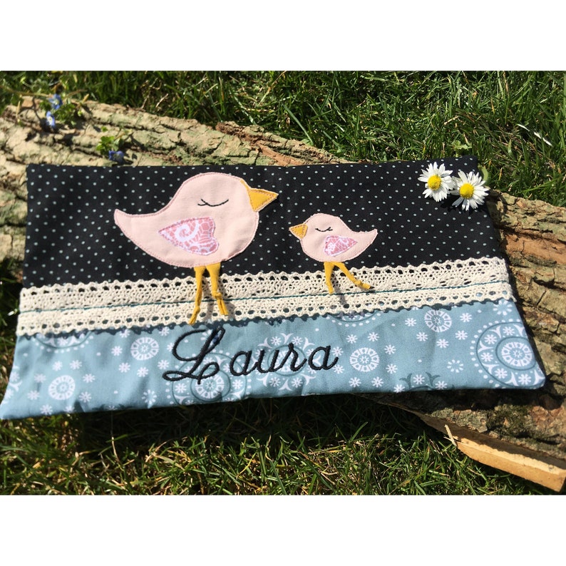 Op de afbeelding: Een zwarte en blauwe stoffen tas met een witte kantversiering. De tas heeft twee geborduurde roze vogels met gele snavels en poten. De tas heeft de naam "Laura" geborduurd in zwart op de voorkant.