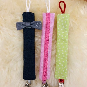 May include: Three fabric baby teethers with clips. The teethers are made of different fabrics and have different patterns. One is navy blue with a gray bow, one is pink with white polka dots, and one is green with white stars.