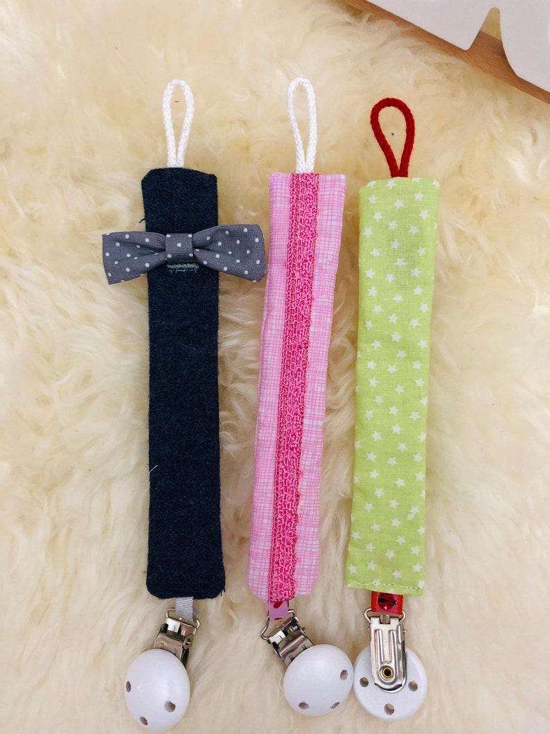 May include: Three fabric baby teethers with clips. The teethers are made of different fabrics and have different patterns. One is navy blue with a gray bow, one is pink with white polka dots, and one is green with white stars.