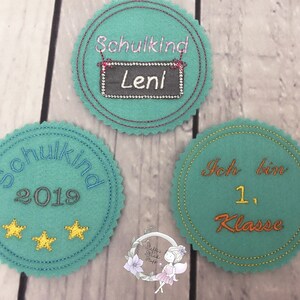 Pode incluir: Tr&ecirc;s c&iacute;rculos de feltro com texto bordado. O primeiro c&iacute;rculo &eacute; azul turquesa com o texto "Schulkind 2019" e duas estrelas douradas. O segundo c&iacute;rculo &eacute; azul turquesa com o texto "Schulkind" e um quadro-negro com o texto "Leni". O terceiro c&iacute;rculo &eacute; azul turquesa com o texto "Ich bin 1. Klasse".