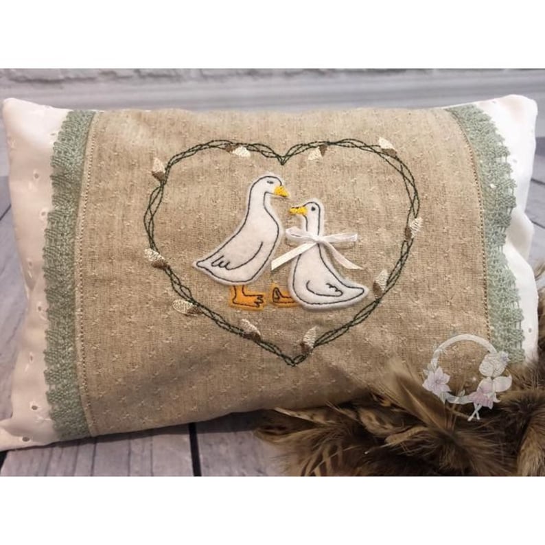 May include: A beige pillow with a green embroidered heart design featuring two white ducks. The pillow has a white lace trim with green accents.