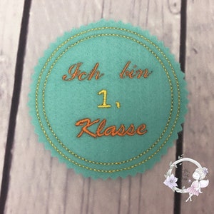 Pode incluir: Um c&iacute;rculo de feltro azul turquesa com uma borda costurada dourada. O c&iacute;rculo tem o texto "Ich bin 1. Klasse" bordado em fio laranja e amarelo.