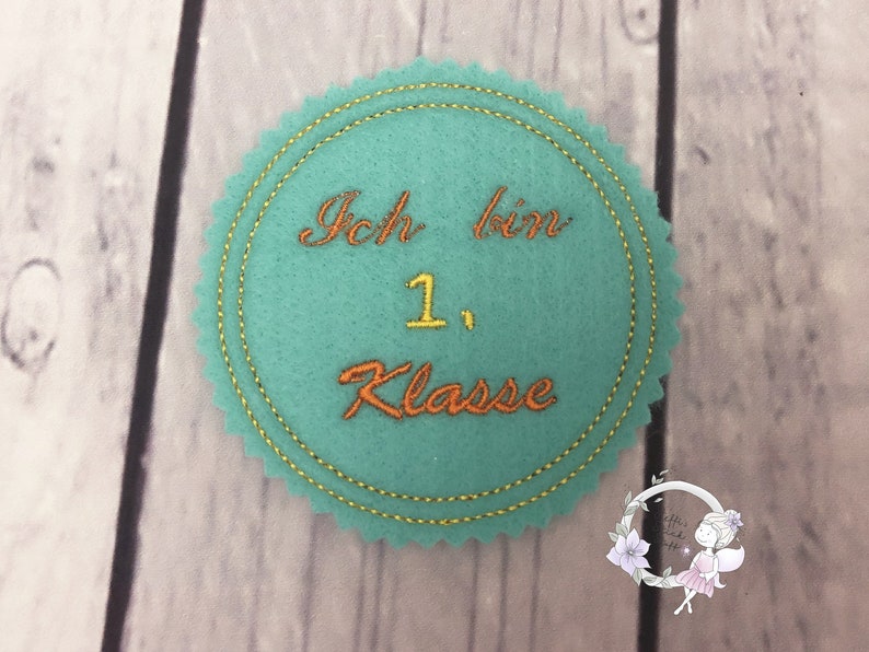 Pode incluir: Um c&iacute;rculo de feltro azul turquesa com uma borda costurada dourada. O c&iacute;rculo tem o texto "Ich bin 1. Klasse" bordado em fio laranja e amarelo.