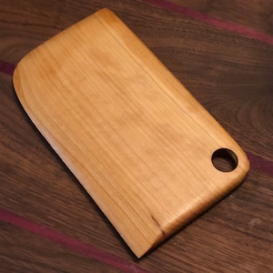Op de afbeelding: Een klein, rechthoekig houten snijplank met een afgeronde rand en een gat in de rechterbovenhoek. De plank is gemaakt van licht hout met een natuurlijke houtnerf.