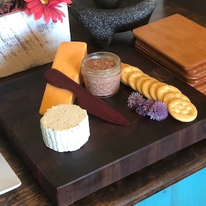 May include: A dark brown wooden cutting board with a variety of snacks, including crackers, cheese, and a small jar of pate. The board is surrounded by purple flowers and a wooden spoon.