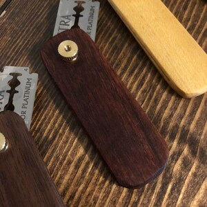 May include: Close-up of a dark brown wooden razor handle with a brass screw. The handle is rectangular with rounded edges. Several silver razor blades are visible in the background, along with a light-colored wooden handle.