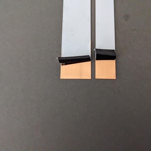 May include: Two flat, white flexible ribbon cables positioned side-by-side. Each cable features a rectangular copper-coloured contact pad at the bottom end, partially covered by a small, black adhesive strip that is peeling away from the surface.