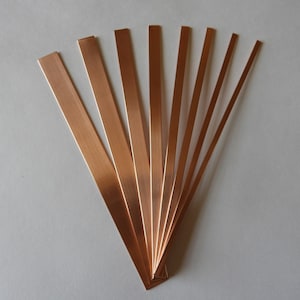 May include: Several copper strips of varying widths are fanned out against a plain, light background. The strips have a polished, metallic appearance. These copper strips, likely for crafting or decorative use, are presented in a visually appealing arrangement.