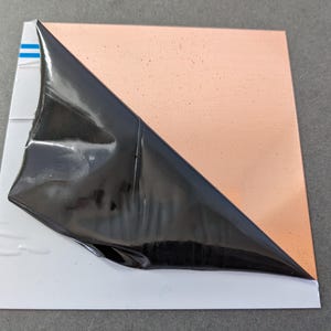 May include: A square copper sheet with a protective black film partially peeled back, revealing the copper surface. The copper has a slightly textured appearance. The sheet is on a white backing with blue stripes.