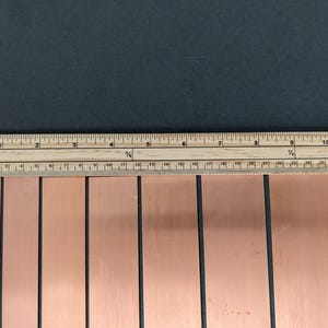 May include: A wooden ruler measuring copper strips. The ruler is marked in inches and fractions of an inch. The copper strips are rectangular and have a reddish-brown colour. The background is a dark grey.