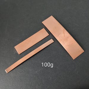 May include: Three rectangular copper strips of varying widths and lengths arranged in a cluster. The metal pieces have a smooth, metallic finish with a reddish-brown hue. The text 100g is printed in white near the bottom of the arrangement.
