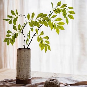 May include: A white ceramic vase with a brown base, holding a branch with green leaves. The vase is sitting on a light brown surface.