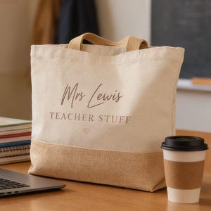 A beige tote bag with the text "Mrs Lewis Teacher Stuff" sits on a wooden desk, accompanied by a laptop, a stack of books, and a coffee cup.