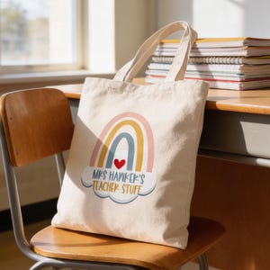May include: A cream-colored tote bag with a rainbow design and the text "Mrs. Hawkers Teacher Stuff." The bag is sitting on a wooden chair in a classroom setting. A desk with books, pencils, and an apple is in the background.