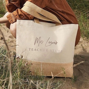 A woman in a brown jacket sits in a field, holding a bag with the text "Mrs Lewis Teacher Stuff" printed on it.