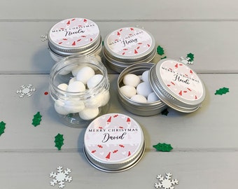 May include: Four small silver tins with white lids, each with a personalized Christmas label featuring a gnome design and the names Nicola, Harry, Heidi, and David. The tins are filled with white candy and scattered on a gray wooden surface with red and green confetti.
