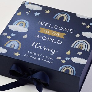 May include: A navy blue gift box with a matching ribbon. The box is decorated with watercolor rainbows, clouds, stars, and raindrops. The text on the box reads "WELCOME TO THE WORLD Harry" with a personalized message below.