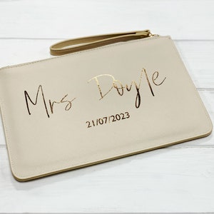 May include: A beige clutch bag with a gold zipper and a gold handle. The bag has the text "Mrs. Doyle" and the date "21/07/2023" printed in gold.