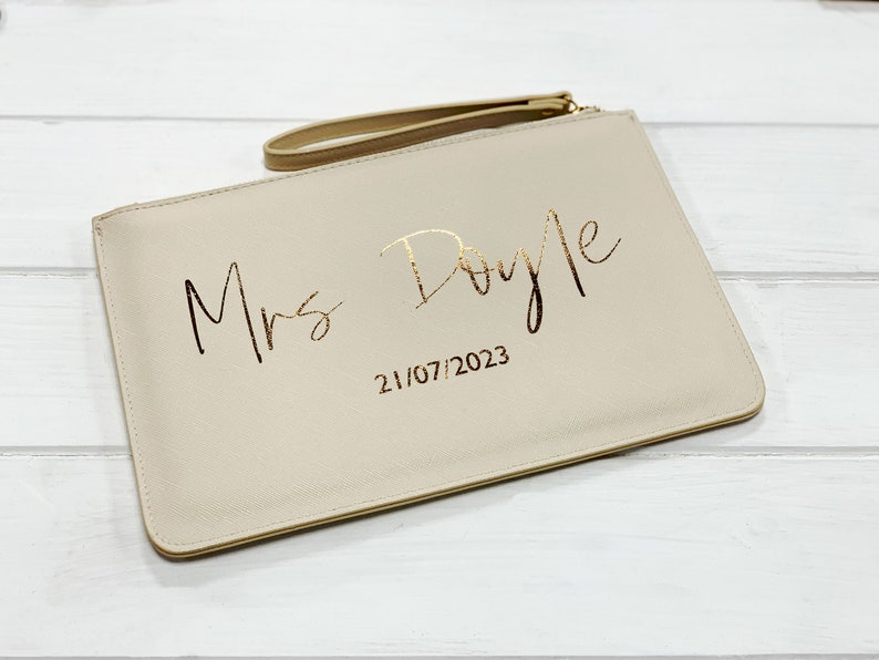 May include: A beige clutch bag with a gold zipper and a gold handle. The bag has the text "Mrs. Doyle" and the date "21/07/2023" printed in gold.
