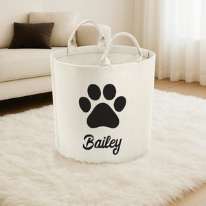 A white tote bag with a black paw print and the name "Biley" printed on it, placed on a white shag rug in a living room.